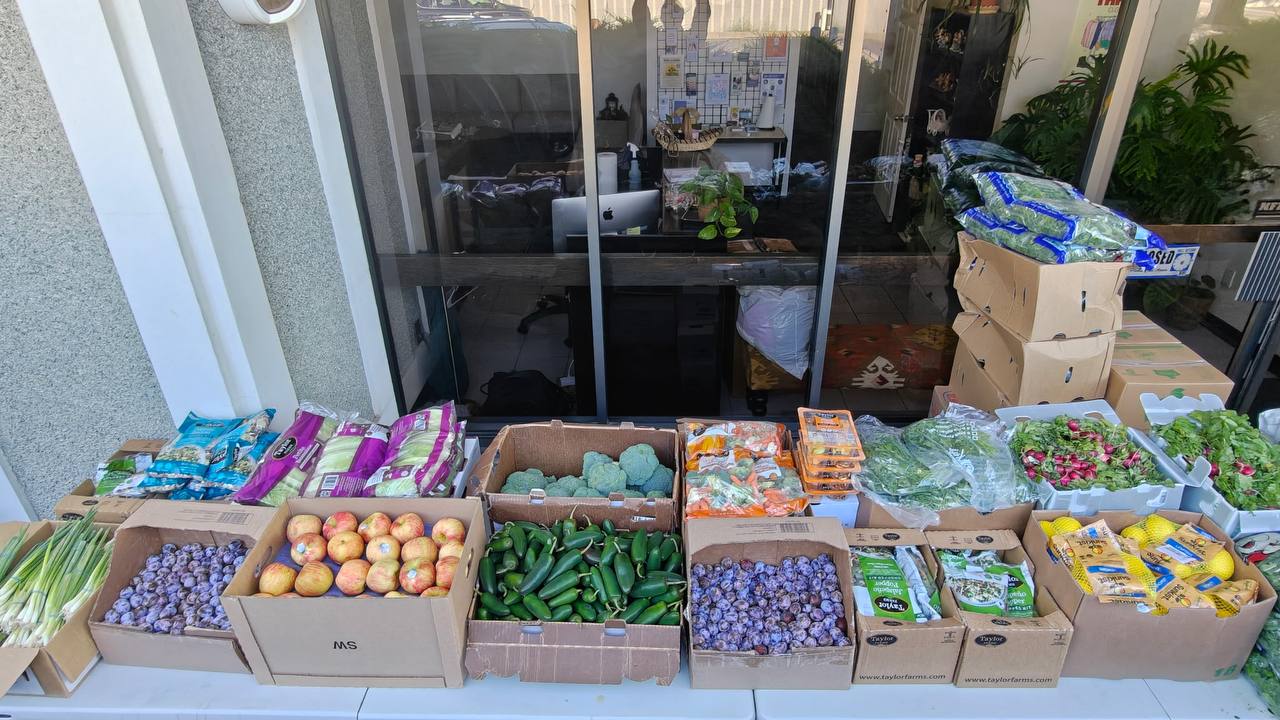 Boxes of fresh produce — plums, apples, peppers, broccoli and greens — set out for Food Bank day outside SSA.